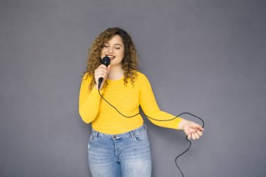 Şirin esmer artı boyutu kadın sarı kazak ve bir nötr gri arka plan üzerinde ayakta kot kıvırcık saçlı. O mocrophone onun ile şarkı şarkı ve mutlu bir şekilde dans elele