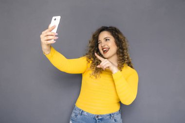 Şirin esmer artı boyutu kadın sarı kazak ve bir nötr gri arka plan üzerinde ayakta kot kıvırcık saçlı. Selfie, kendisini cep telefonu fotoğraf çekimi yapar. Kopya alanı