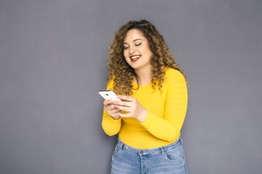 Şirin esmer artı boyutu kadın sarı kazak ve bir nötr gri arka plan üzerinde ayakta kot kıvırcık saçlı. O cep telefonu sms, ne okudu hayret yazarak sohbet