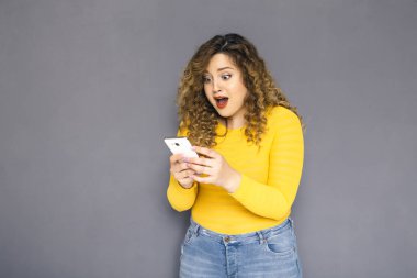Şirin esmer artı boyutu kadın sarı kazak ve bir nötr gri arka plan üzerinde ayakta kot kıvırcık saçlı. O cep telefonu sms, ne okudu hayret yazarak sohbet