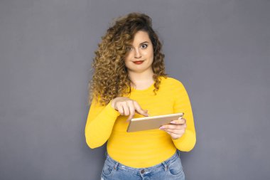 Şirin esmer artı boyutu kadın sarı kazak ve bir nötr gri arka plan üzerinde ayakta kot kıvırcık saçlı. O tablet kullanmayı deneyin ve nasıl ile çalışır bilmiyorum