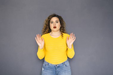 Şirin esmer artı boyutu kadın sarı kazak ve bir nötr gri arka plan üzerinde ayakta kot kıvırcık saçlı. O hayran, bazı sürpriz hakkında şok