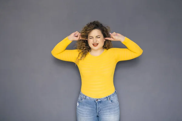 Cute brunette plus size woman with curly hair in yellow sweater and jeans standing on a neutral grey background. She pretends she not listening, stucking fingers on her ears