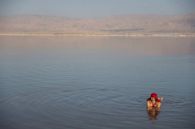 Kızıl mayo ve kırmızı şapkayla İsrail 'de ölü denizde yüzen bir kadın. Mükemmel mavi su ve gökyüzü. Kopyalama alanı olan yaz manzaralı resim.