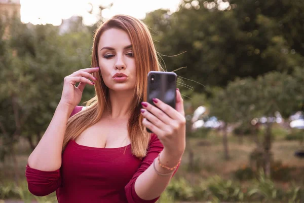 Caucasian beautiful woman in casual but sexy outfit standing in park, outdoor, holding her phone in her arms and make dumb selfie. Summer evening weather. Space for text.