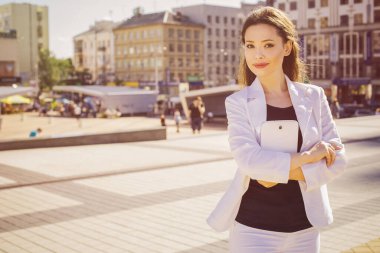 Beyaz takım elbiseli güzel esmer iş kadını açık havada elinde tabletle çalışıyor. Boşluğu kopyala