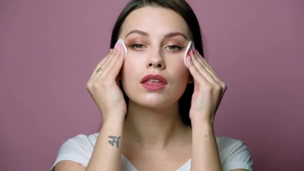 Sexy brunette caucasian woman in neutral clothes standing on a pink background. She clean her face using sponges, getting rid of make up. Cosmetic concept — Stock Video