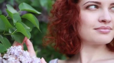 Yazlık elbiseli, kızıl saçlı beyaz kadın parkta rahatlıyor, leylak çiçekleri kokluyor ve mutlu bir şekilde gülümsüyor. Güneşli bir bahar gününde vesikalık portre