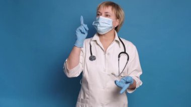 Beyaz önlüklü, mavi arka planda, Kafkasyalı, yaşlı bir doktor. Steteskop ve eldiven takıyor ve parmaklarını işaret ediyor.