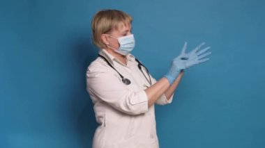 Beyaz önlüklü, mavi arka planda, Kafkasyalı, yaşlı bir doktor. Steteskop takıyor ve eldiven takıyor.