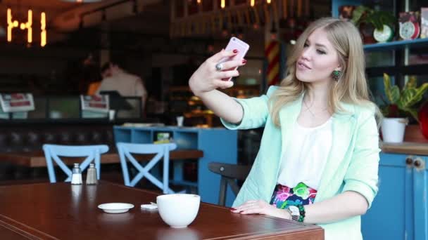 Γυναίκα το καλοκαίρι έξυπνη περιστασιακή outfint κάθεται σε μια βεράντα καφέ, πίνοντας καφέ και κάνοντας selfie στο τηλέφωνό της. Σέξι χαριτωμένο χαμογελαστό ξανθιά θέτουν και φλερτ με την κάμερα — Αρχείο Βίντεο