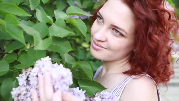 Mujer caucásica linda pelirroja en vestido de verano relajante en el parque, oliendo flores lila y sonriendo felizmente. Retrato de la cabeza en un soleado día de primavera — Vídeos de Stock