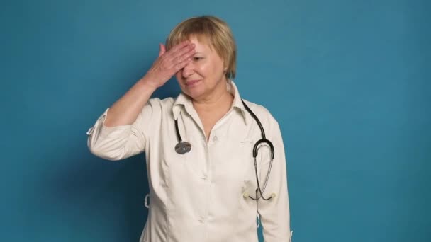 Γηραιή καυκάσια γιατρός με λευκό παλτό σε μπλε φόντο. Φοράει στηθοσκόπιο και κάνει απογοητευμένες χειρονομίες. — Αρχείο Βίντεο