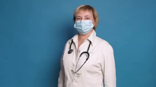青を背景に白衣を着た老白人医師の女性。彼女は聴診器とマスクを着用し、親指を作る/ジェスチャーのように — ストック動画