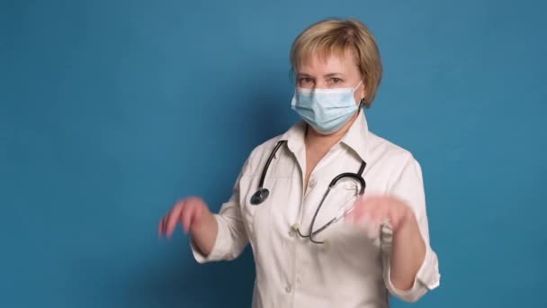 Starší bělošská doktorka v bílém plášti na modrém pozadí. Nosí stetoskop a nasazuje si masku. — Stock video