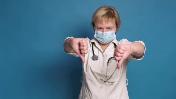 Oudere blanke dokter dame in witte jas op blauwe achtergrond. Ze draagt stethoscoop en maakt duimen naar beneden / afkeer van gebaren — Stockvideo