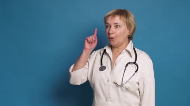 Donna anziana medico caucasico in cappotto bianco su sfondo blu. Indossa lo stetoscopio e punta le dita questo gesto Idea — Video Stock