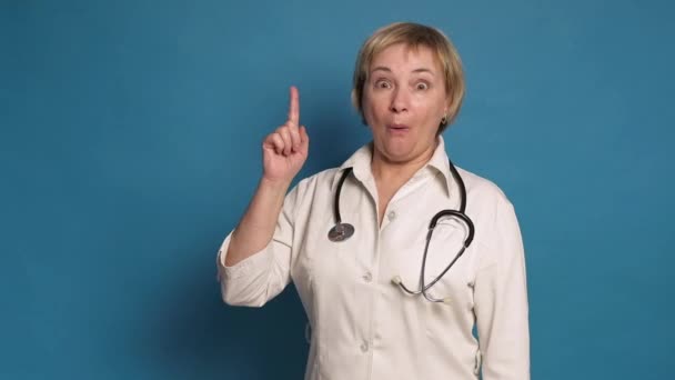 Starší bělošská doktorka v bílém plášti na modrém pozadí. Nosí stetoskop a ukazuje na něj prstem. — Stock video