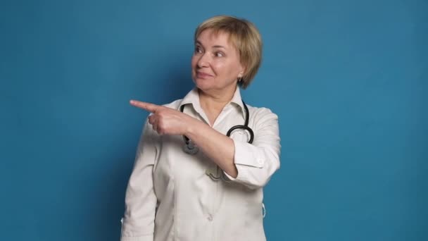 Anciana doctora caucásica en bata blanca sobre fondo azul. Ella usa estetoscopio y señala sus dedos — Vídeos de Stock