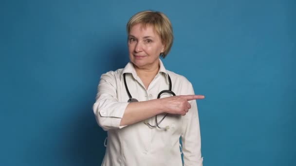 青を背景に白衣を着た老白人医師の女性。彼女は聴診器をつけて指を指差す — ストック動画
