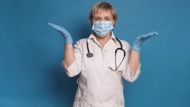 Eldery kaukasische Ärztin in weißem Mantel auf blauem Hintergrund. Sie trägt Stethoskop und macht frustrierte Gesten — Stockvideo