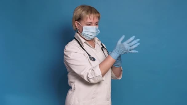 Eldery caucasian doctor lady in white coat on blue background. She wear stethoscope and putting on gloves — Stock Video