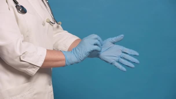 Starší bělošská doktorka v bílém plášti na modrém pozadí. Zblízka ruce, nasazování rukavic — Stock video