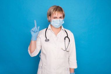 Beyaz önlüklü, yaşlı, beyaz saçlı, mavi arka planda steteskoplu bir doktor. Duygusal portreler: parmaklarını gösteriyor..