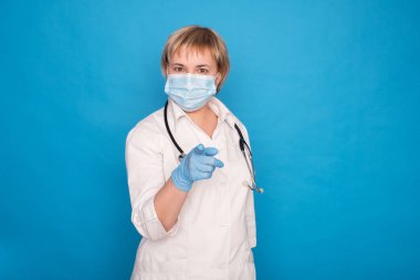 Beyaz önlüklü, yaşlı, beyaz saçlı, mavi arka planda steteskoplu bir doktor. Duygusal portreler: parmaklarını gösteriyor..