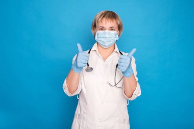 Beyaz önlüklü, yaşlı, beyaz saçlı, mavi arka planda steteskoplu bir doktor. Duygusal portreler, baş parmaklar..