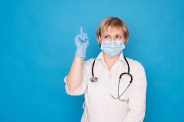 Beyaz önlüklü, yaşlı, beyaz saçlı, mavi arka planda steteskoplu bir doktor. Duygusal portreler: parmaklarını gösteriyor..