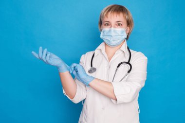 Beyaz önlüklü, yaşlı, beyaz saçlı, mavi arka planda steteskoplu bir doktor. Eldiven takıyor..
