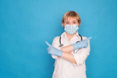 Beyaz önlüklü, yaşlı, beyaz saçlı, mavi arka planda steteskoplu bir doktor. Duygusal portreler: parmaklarını gösteriyor..