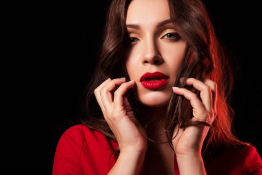 Seksi esmer kadın, siyah arka planda kırmızı şık elbiseler içinde duruyor. Çekici bir şekilde poz veriyor ve gülümsüyor.