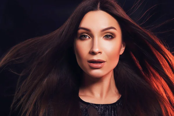 Close up portrait of a woman with flawless skin and evening make up. She wears black dress with lace and stands on black background with red backlight. Her hair fly on a wind. Sexy, sensual posing.