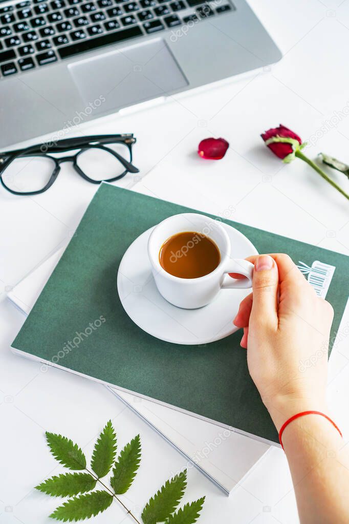a female hand holds a coffee mug, stands on a magazine, next to it are glasses and a red rose. Worth a laptop