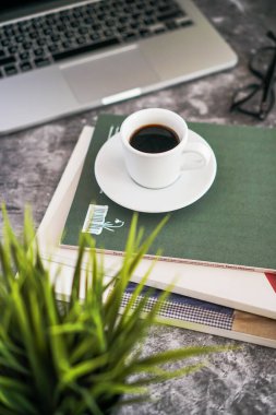 Bir kahve fincanı dergilerin üzerinde duruyor, sonra bir dizüstü bilgisayar ve bir çiçek.