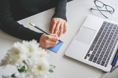 Kız not defterine not alıyor. Yanında dizüstü bilgisayar ve vazo çiçek var.