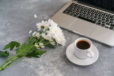 Kahve fincanı, dizüstü bilgisayar ve beyaz kasımpatılar gri arka planda masanın üstünde.