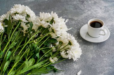 Masanın üzerinde bir fincan kahve ve yanında da beyaz kasımpatılar var.