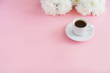 Bir kahve fincanı pembe bir arka planda durur, yanında kasımpatı çiçekleri vardır.