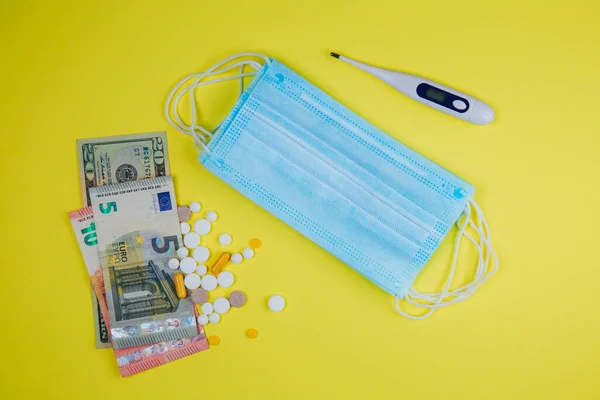 Medical mask and money, thermometer and pills on a yellow background.