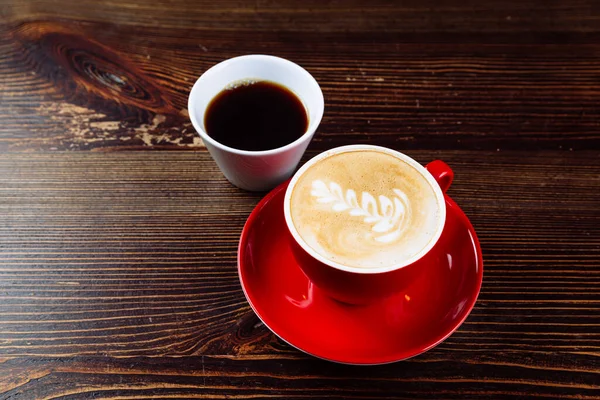 Café en una taza roja con leche y latte arte y café en una taza blanca sobre una mesa —  Fotos de Stock