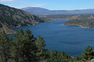 Ön planda rezervuar ve ağaçlarla doğal manzara, arka planda dağlar ve yoğun mavi gökyüzü, El Atazar rezervuarı, Madrid, İspanya