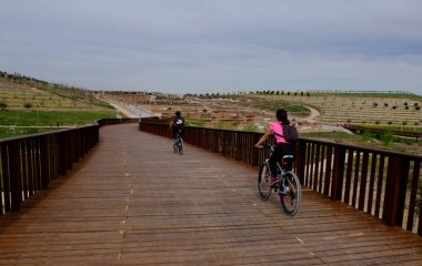 Bisikletçiler, arka planda parkı ve güzel beyaz mavi gökyüzü olan ahşap bir yürüyüş yolu boyunca yürüyorlar, Valdebebas Park, Madrid, İspanya