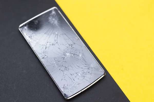 A broken phone on yellow and black background. Crushed device with broken screen representing an accident. Textured screen with damage. Dark glass of a screen.