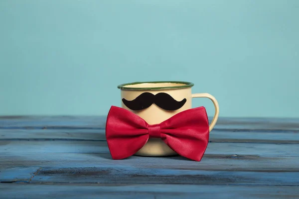 Coffee cup with mustache and red bow tie on blue and vintage wooden background