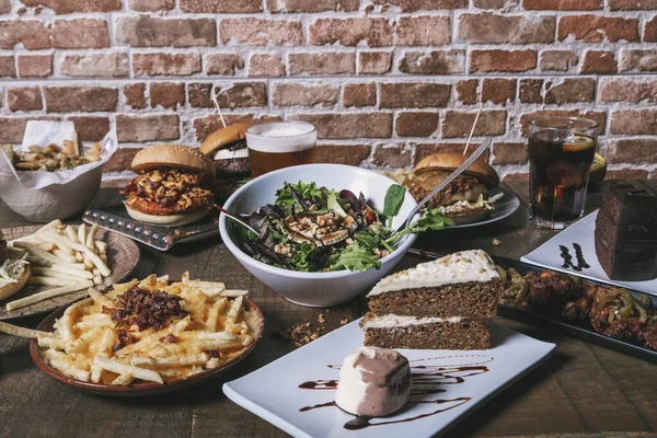 Hamburgerler, patates kızartmaları, salata, içecekler, pasta ve ahşap masada tatlılar..