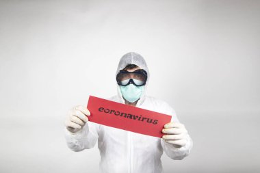 Cerrahi maskeli, gözlüklü, beyaz koruyucu giysili ve üzerinde beyaz arka planda izole edilmiş Coronavirus yazan kırmızı tabelalı bir adam. Yeni salgın yeni hızlı yayılan koronavirüs 2019-ncov. Tıbbi grip virüsü konsepti.