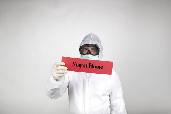 Man in surgical mask with white protective suit and a red sign with the word stay home isolated in studio on white background. New coronavirus pandemic epidemic.